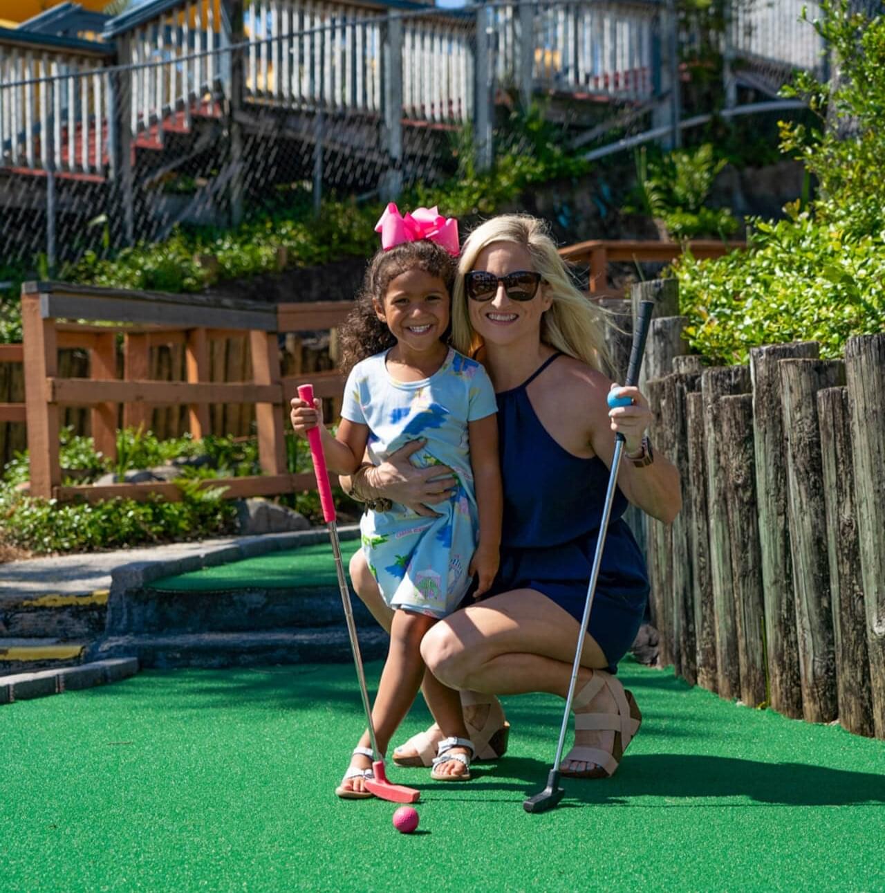 Mother and daughter pose with mini golf clubs smiling on mini golf course